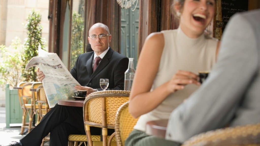 A serious man in a suit reads a newspaper at a cafe while a woman laughs in the foreground.