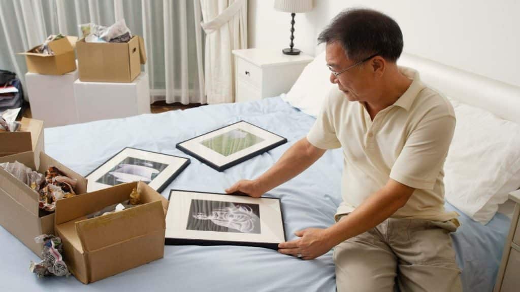 A man sits on a bed, looking at framed pictures surrounded by moving boxes.