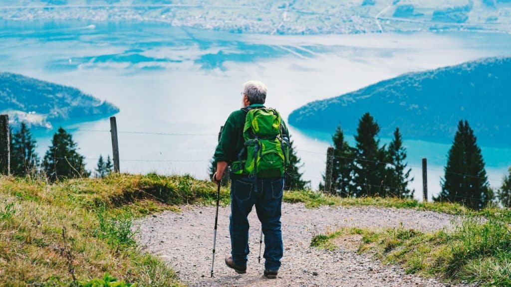 An older man with a green backpack hikes up a scenic mountain path overlooking a large lake.