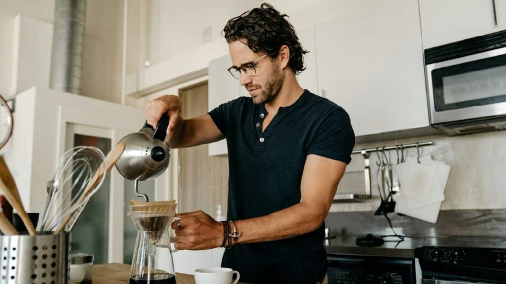 A focused man with glasses pours hot water over a pour-over coffee maker in a bright kitchen.