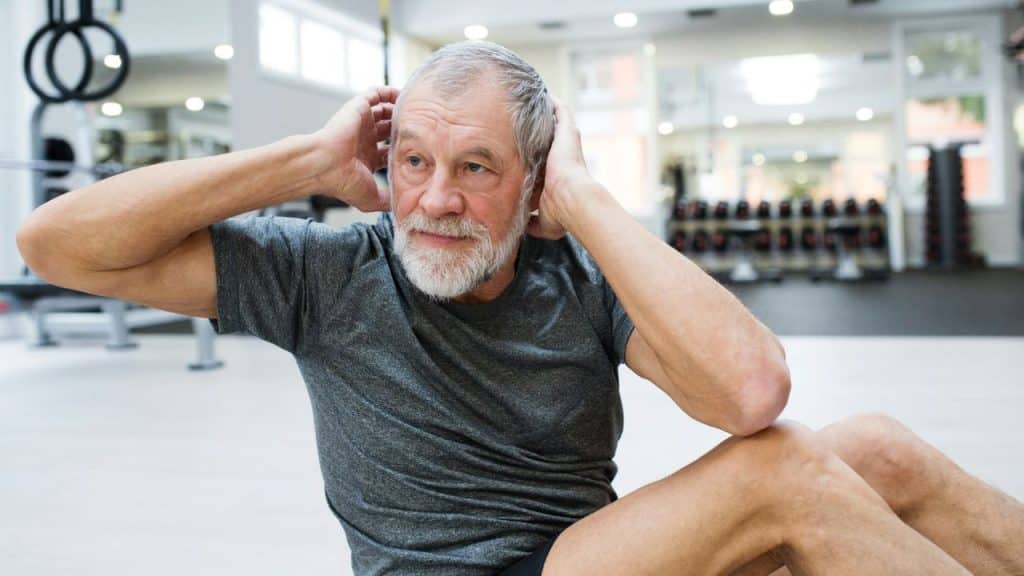 A determined, gray-haired man with a beard does crunches on the floor of a gym.