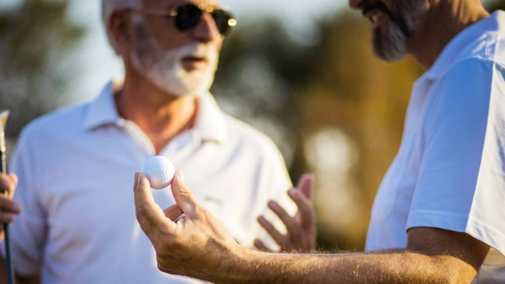 Two senior men in white shirts stand on a golf course, one holding a golf ball.