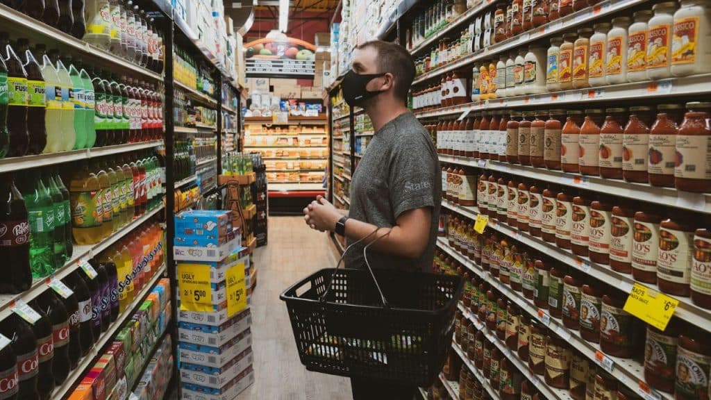 A man wearing a face mask carries a basket while shopping in a grocery store aisle.