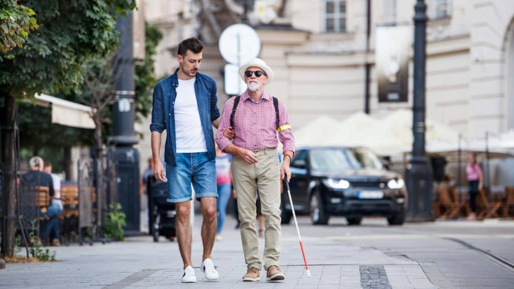 A younger man guides an older man with a white cane along a city sidewalk.