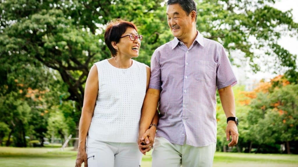 A couple walks hand-in-hand in a park, smiling at each other on a sunny day.