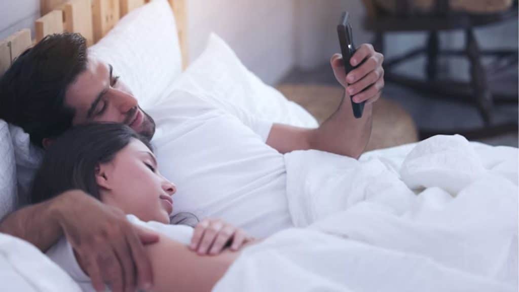 A man watching TV in bed while his partner tries to sleep