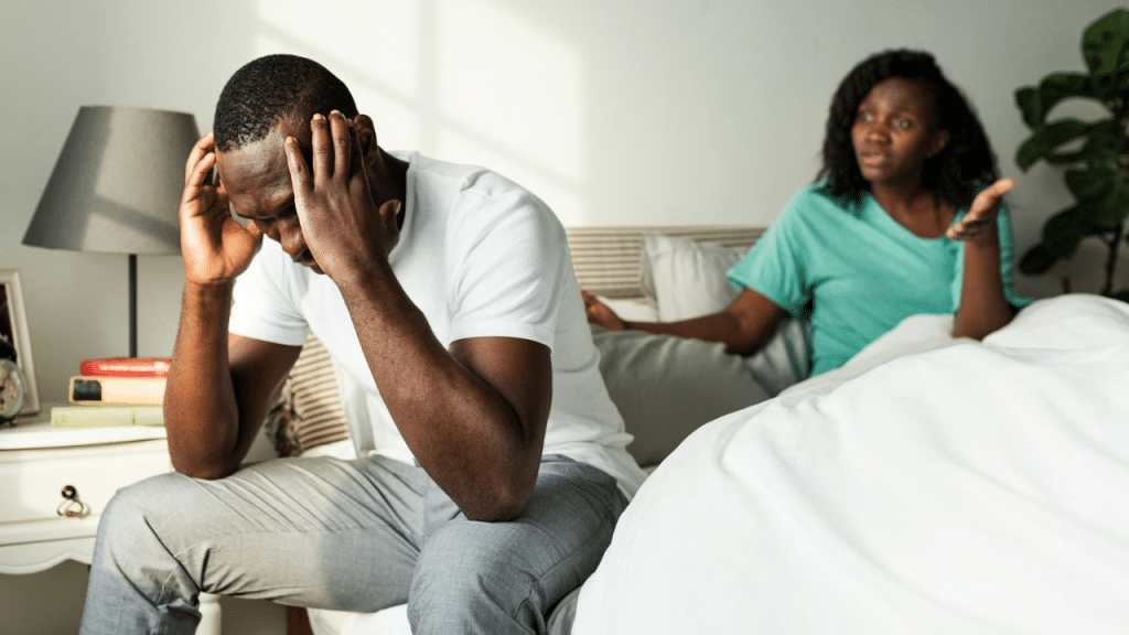A distressed man sits with his head in his hands while a woman argues behind him.