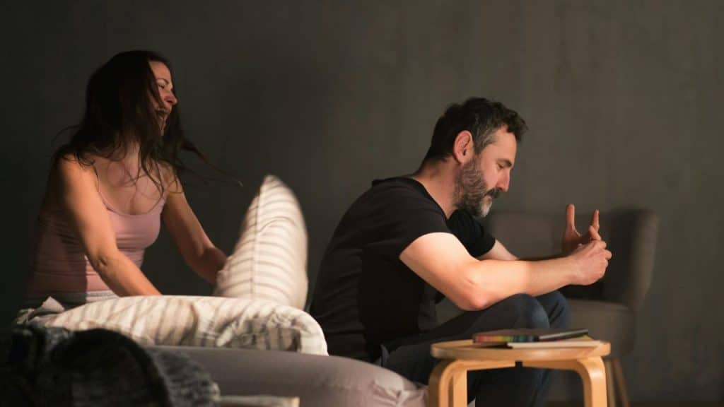 Woman in tank top playfully swings a pillow at a seated, bearded man.