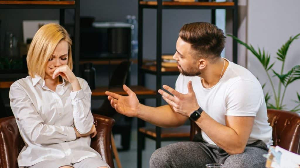 Man in white shirt gestures toward a woman in white who is crying and covering her face.