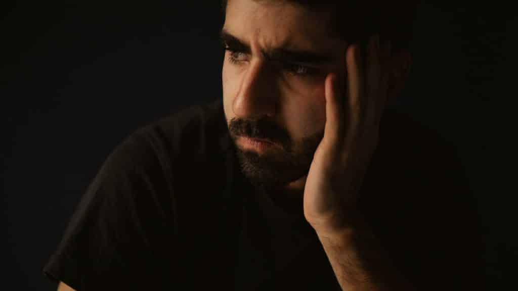 Close-up of a bearded man in a dark shirt with a furrowed brow, resting his head on his hand.