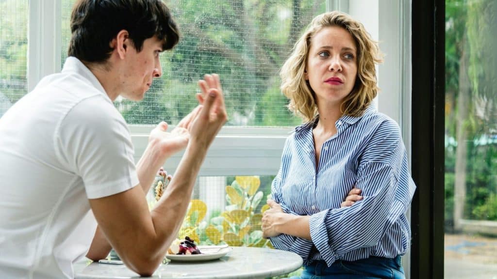Woman with crossed arms and a serious expression watches a man gesturing at a table.