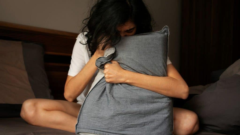 A person with long dark hair sits on a bed, hugging a dark grey pillow to their chest and face.