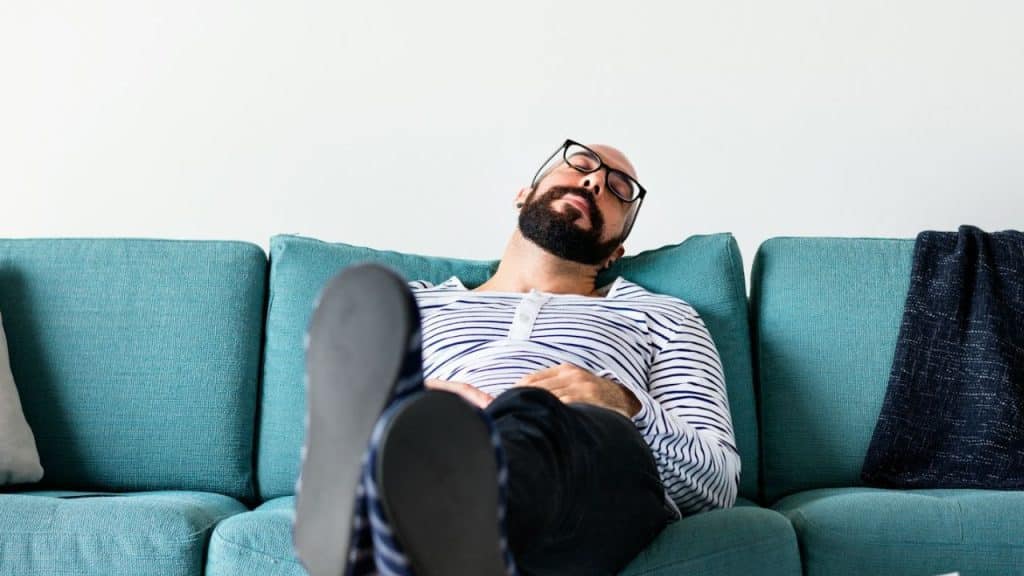 A bald, bearded man wearing glasses and a striped shirt is lying on a turquoise sofa with his eyes closed and feet up.
