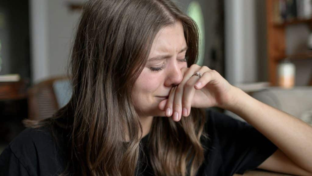 Close-up of a young woman with long brown hair, eyes closed, and her hand covering her mouth.