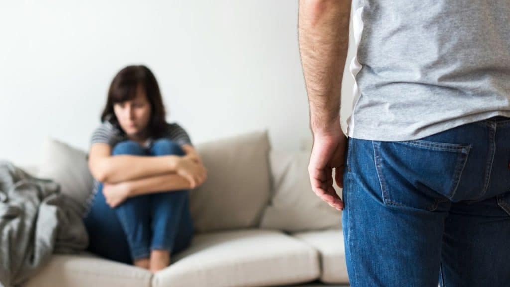A woman sits dejectedly on a sofa while a man in jeans stands with his back to the camera.