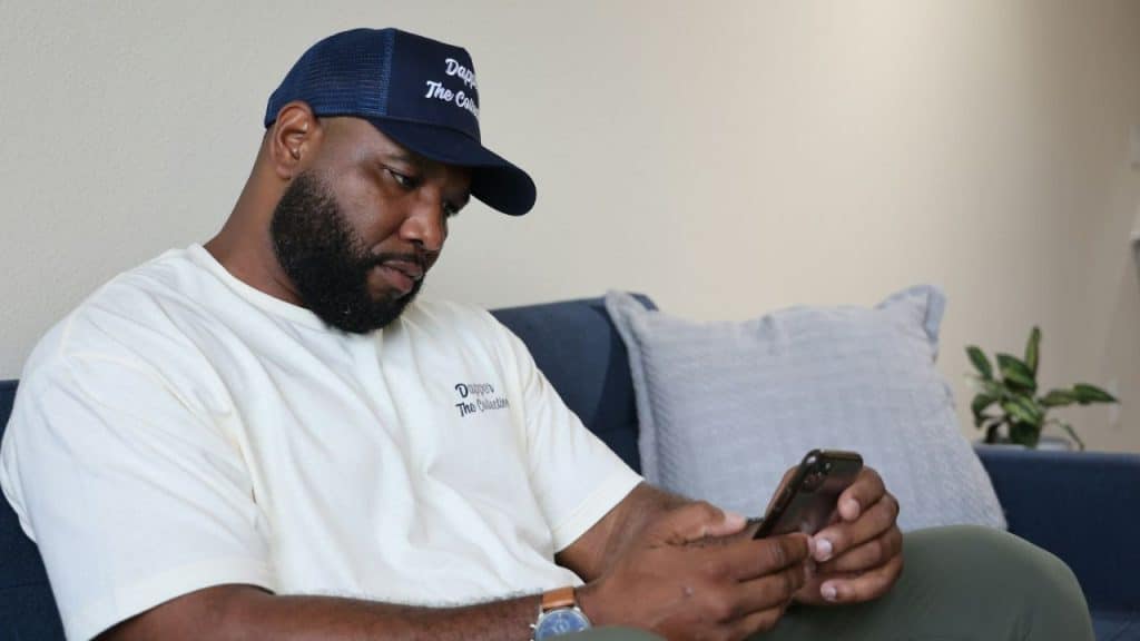 A bearded man wearing a navy blue cap and a light t-shirt is sitting on a sofa looking intently at his phone.