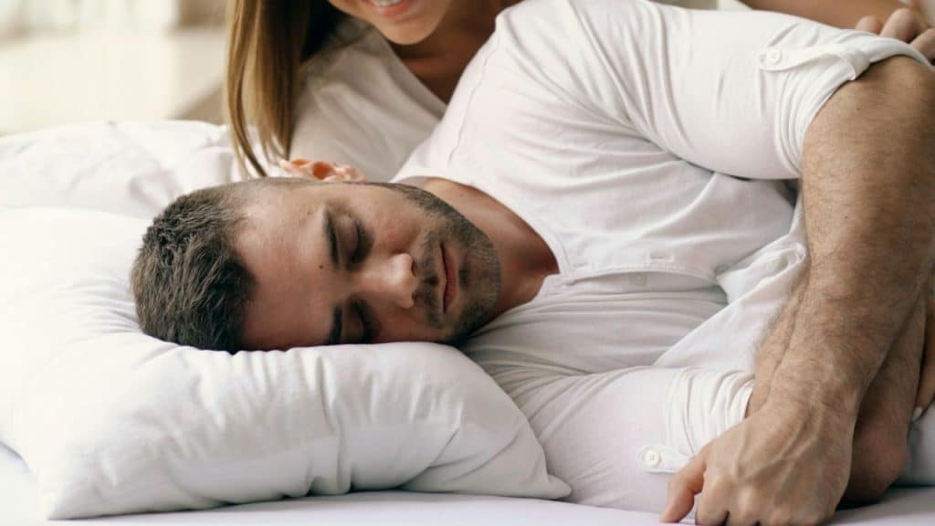 A man with his eyes closed is lying on his side on a white bed, being embraced by a woman behind him.