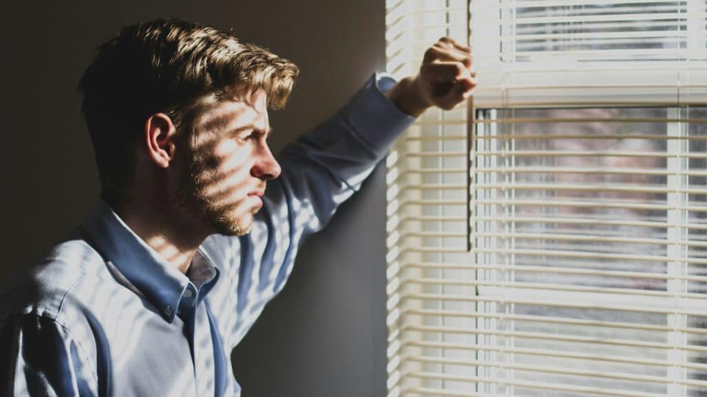 A man in a blue button-down shirt is looking out of a window, pulling the blinds open.
