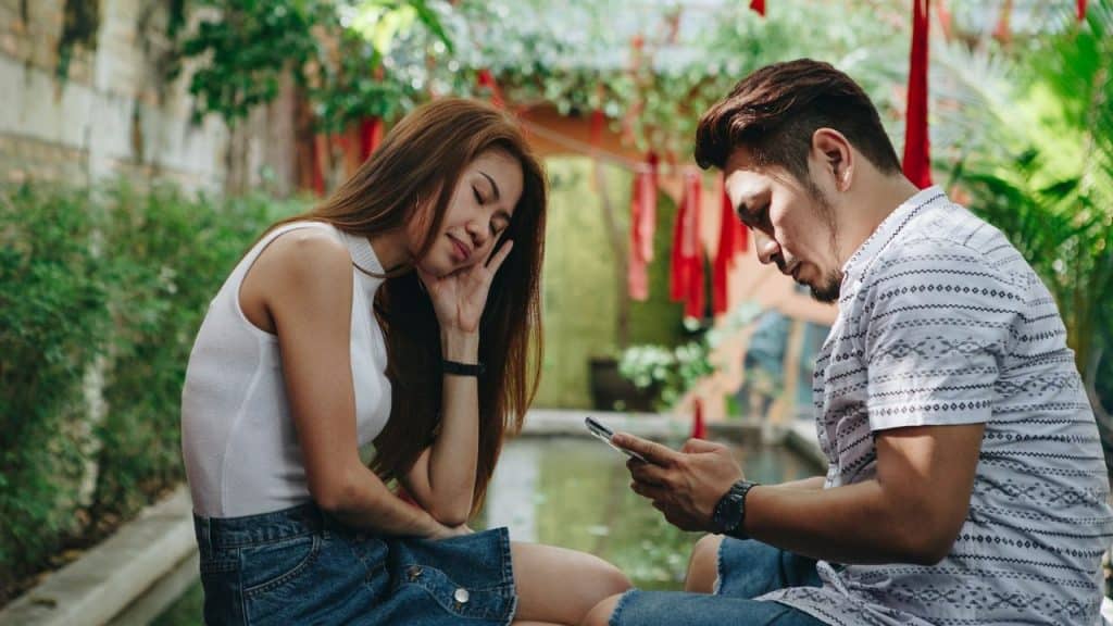 A couple sits outside; the woman is resting her head on her hand, looking bored, and the man is engrossed in his phone.