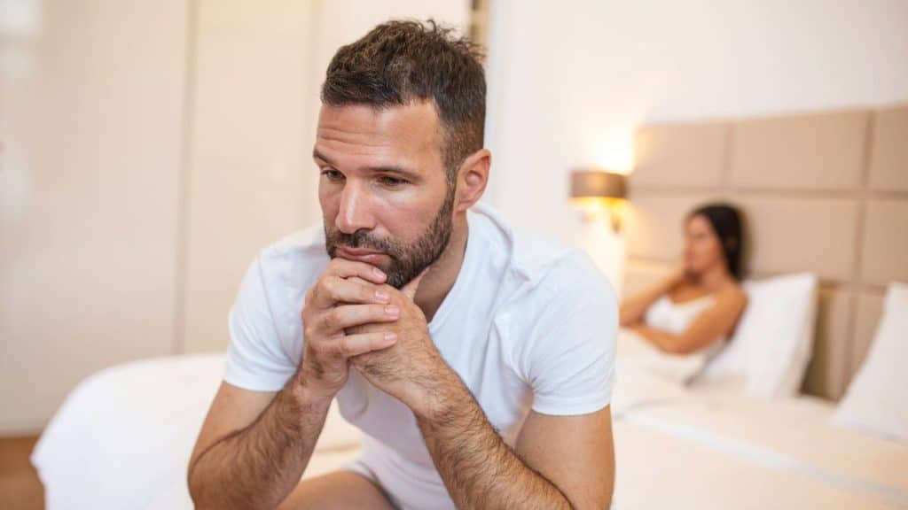 A thoughtful, sad-looking man sits on a bed, with a woman out of focus behind him.