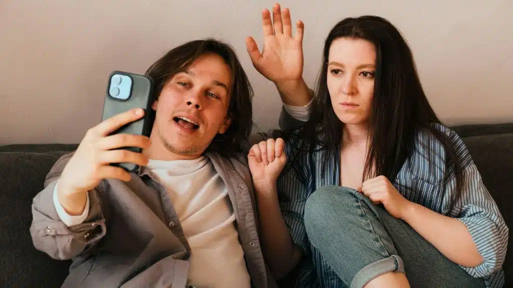 A cheerful man records a video with a phone while a woman waves with an annoyed expression.
