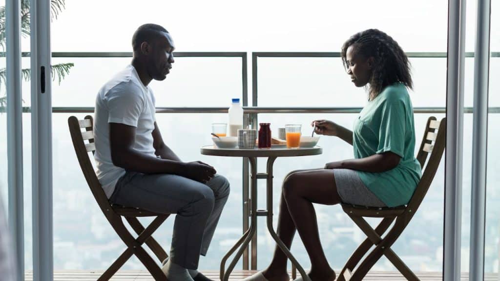 A serious-looking couple sits silently across from each other at a small table on a balcony.