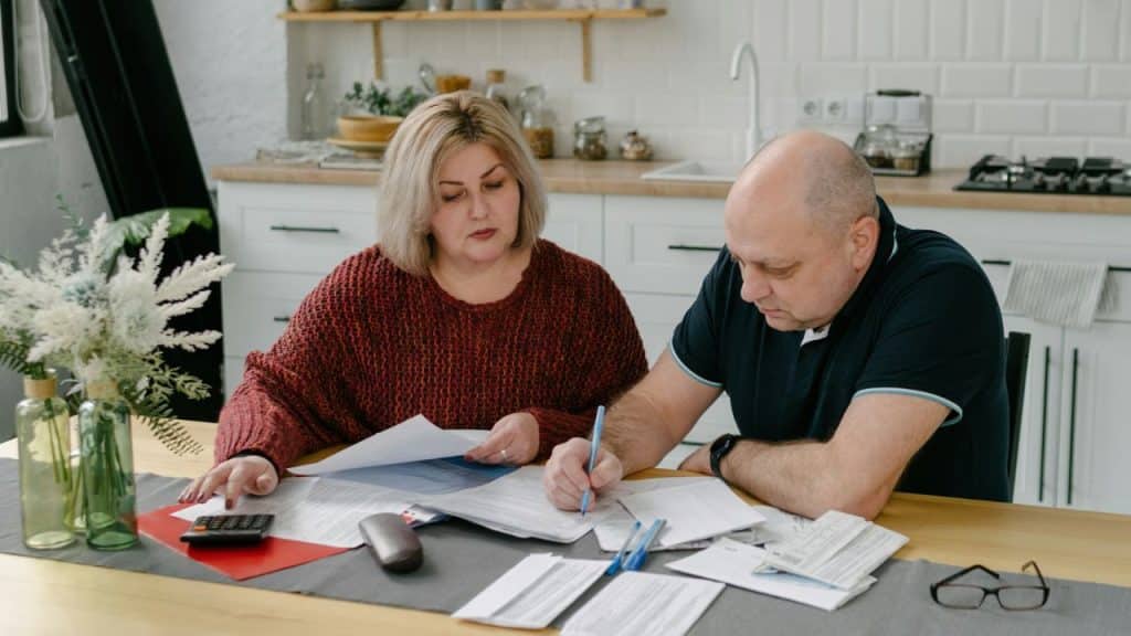 A focused middle-aged couple works together on paperwork and bills at a kitchen table.