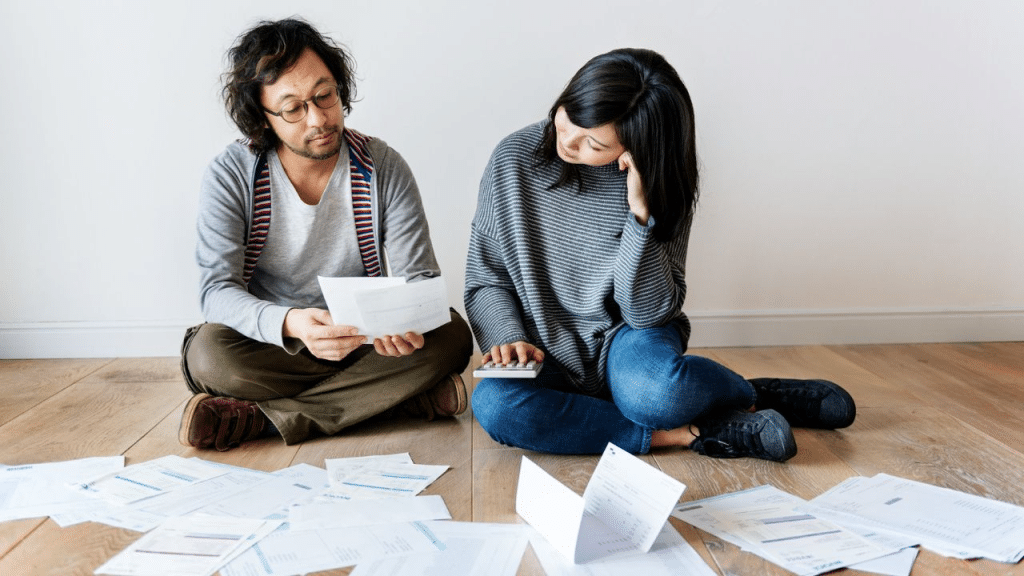 A serious couple sits cross-legged on the floor surrounded by bills and a calculator.
