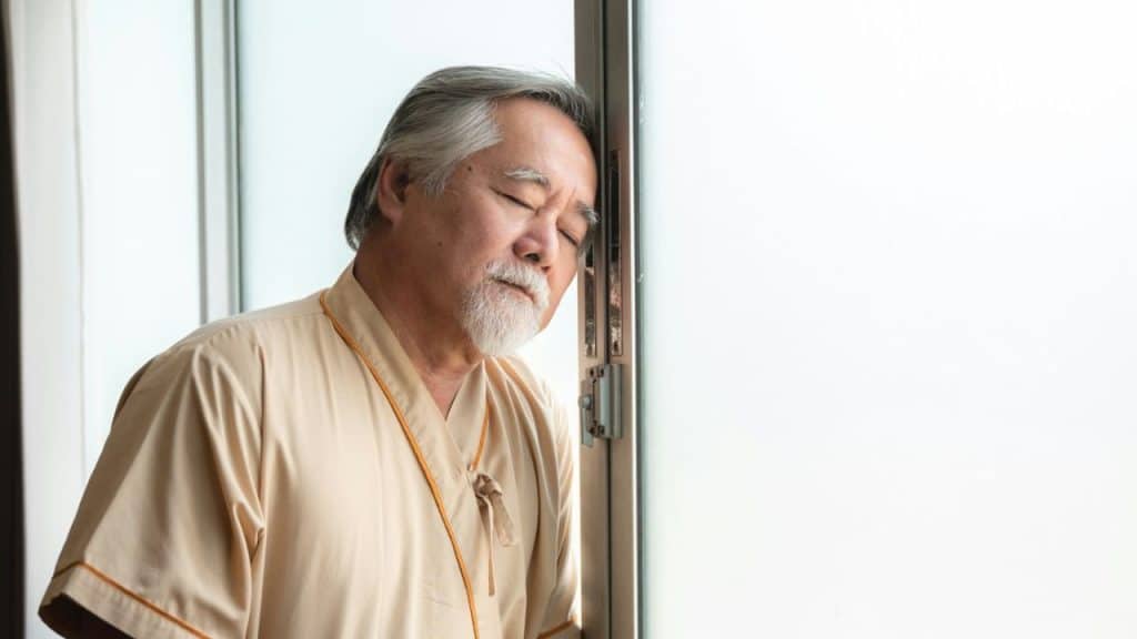 An older man with his eyes closed is leaning his head against a window frame in a brightly lit room.