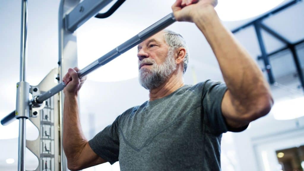 An older man with a grey beard is performing an overhead lift on a **weight machine** in a gym.