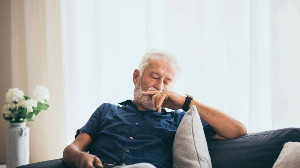 An older man with a grey beard is sitting on a sofa, eyes closed, with his hand near his mouth.