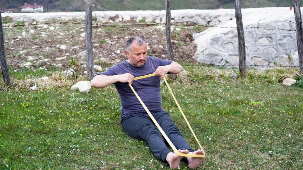 A man sitting outdoors on the grass is performing a stretching exercise with a long yellow resistance band.