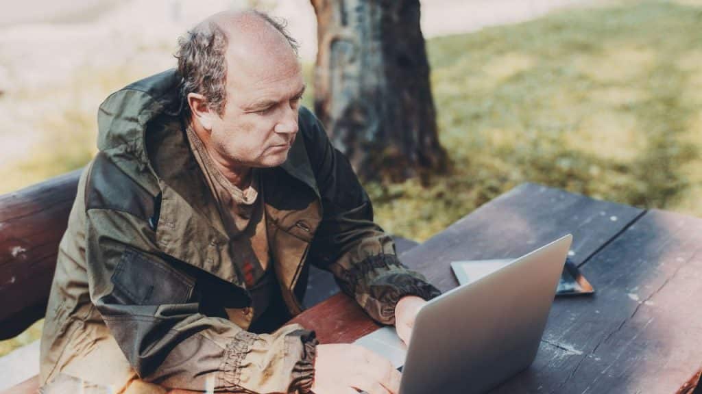 A bald man wearing a green jacket is sitting outdoors at a wooden table using a laptop.