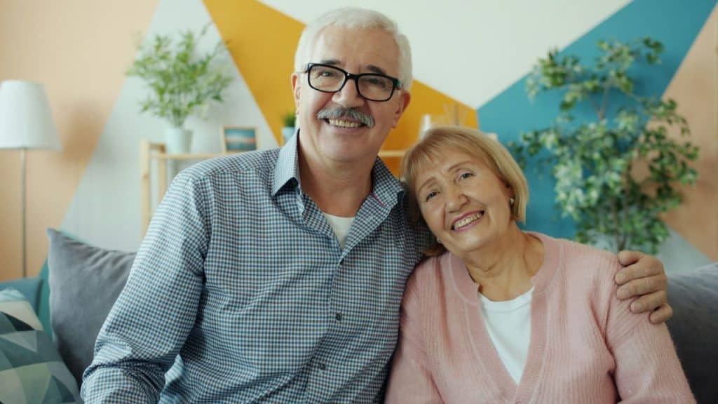 A happy older couple smiles at the camera; the man wears glasses, and the woman leans her head on his shoulder.