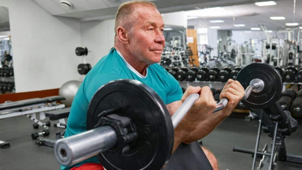An older man in a teal shirt is performing a bicep curl with a barbell in a gym.