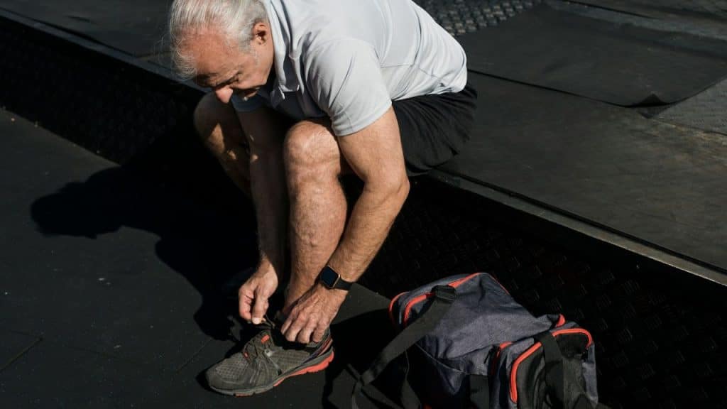 An older man is sitting down, tying his athletic shoe laces next to a duffel bag.
