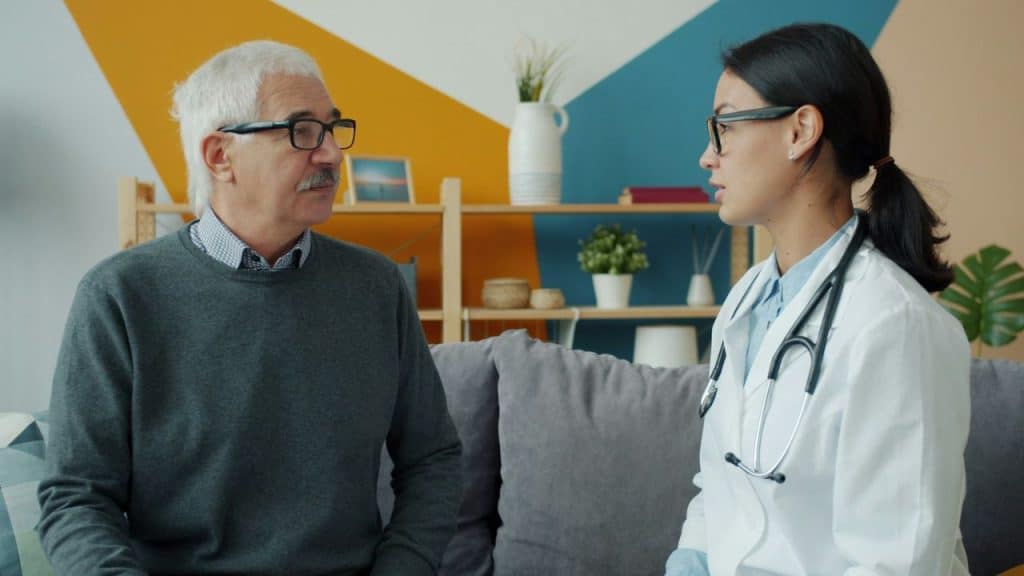 An older man in glasses and a grey sweater is sitting on a sofa and listening to a doctor in a white coat.