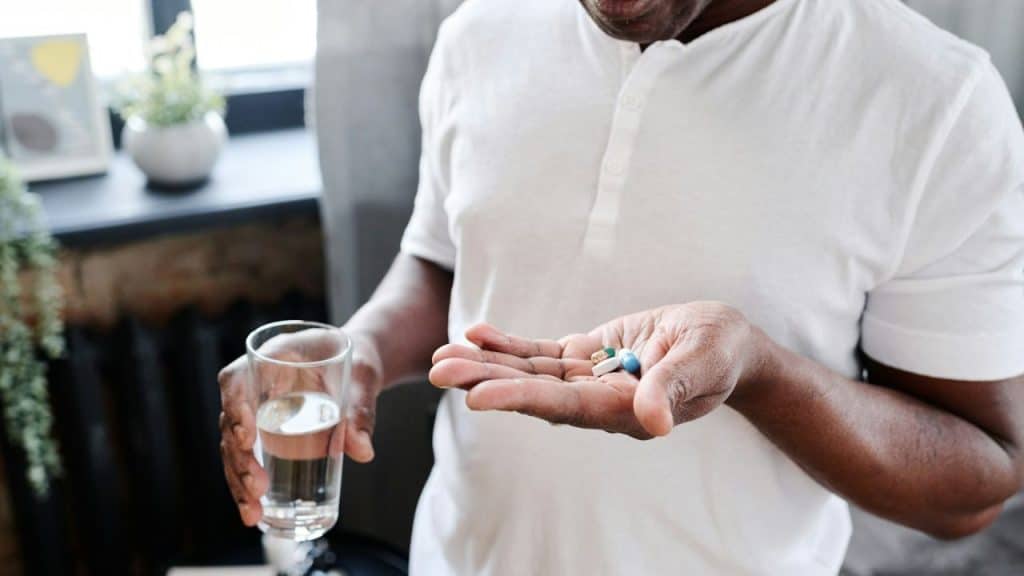 A person in a white shirt holds a small handful of colored pills in one hand and a glass of water in the other.