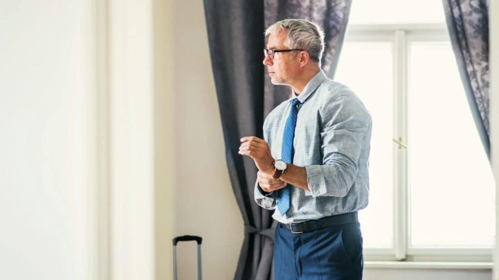 A mature man in a dress shirt, tie, and glasses is adjusting his wristwatch in front of a window.