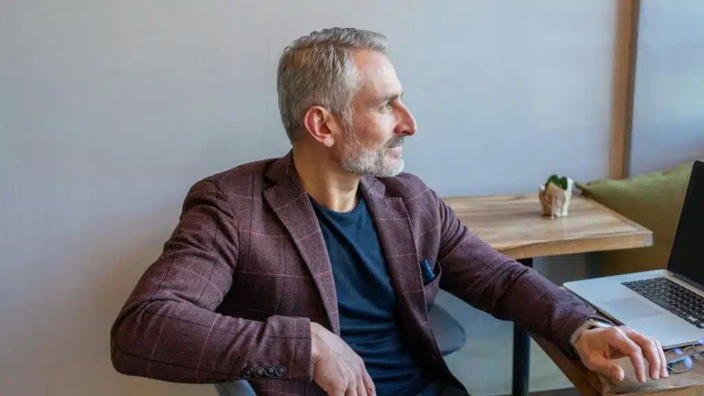 A middle-aged man with gray hair and a beard is sitting at a wooden table with a laptop, looking to the side.