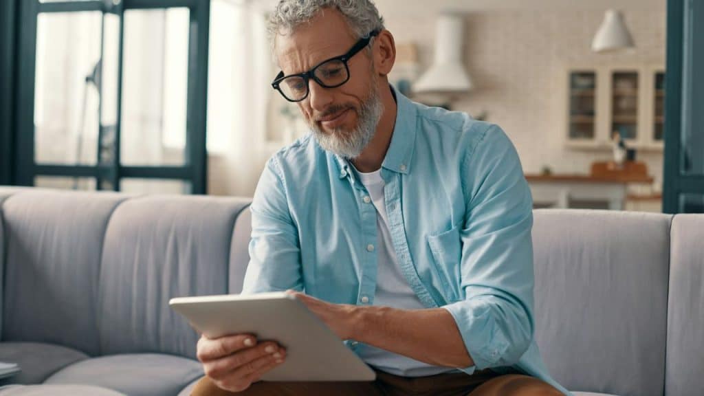 A middle-aged man with gray hair, a beard, and glasses is sitting on a sofa looking at a tablet.
