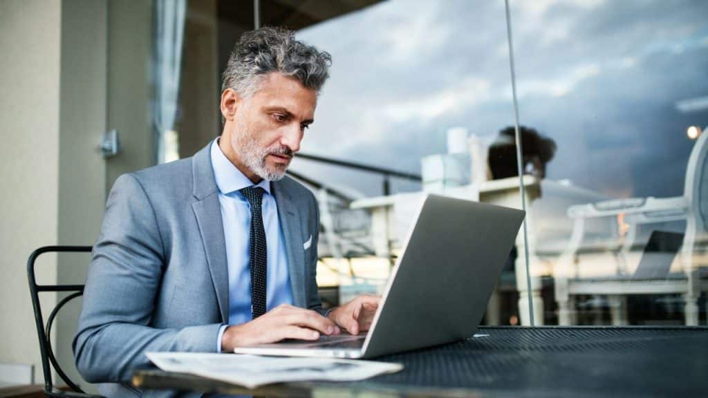 A well-dressed man with grey hair and a beard is intensely focused on his laptop outdoors.