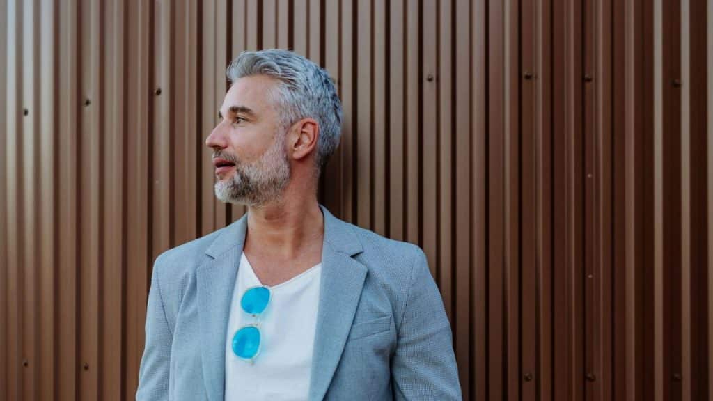 A handsome man with gray hair and a beard, wearing a blazer, stands against a corrugated brown wall.