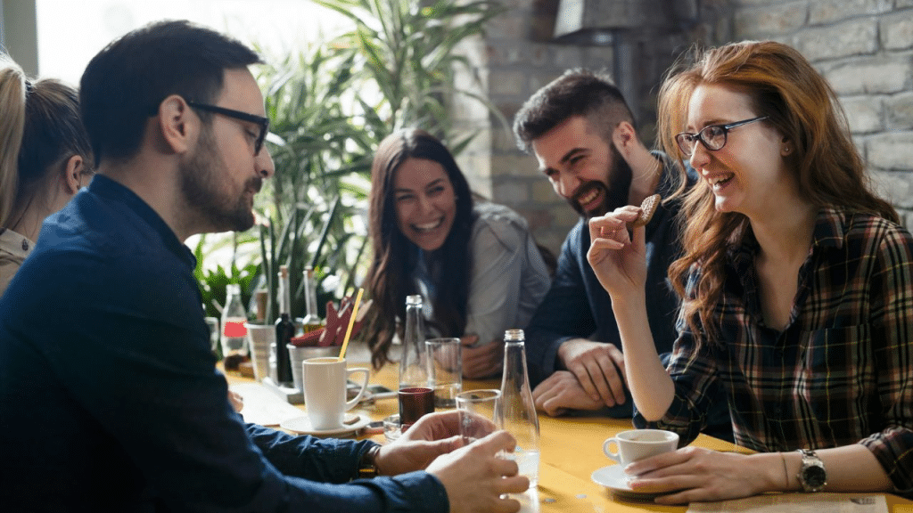 Four smiling friends enjoy coffee and conversation around a bright yellow table at a restaurant.