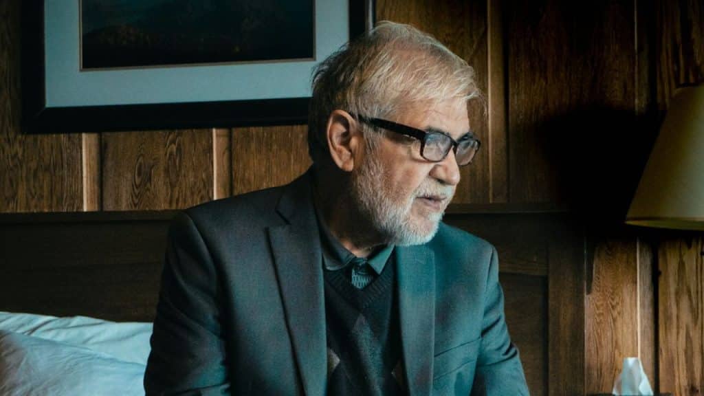A middle-aged man with a white beard, wearing glasses, a grey blazer, and a patterned sweater, is looking to the side.