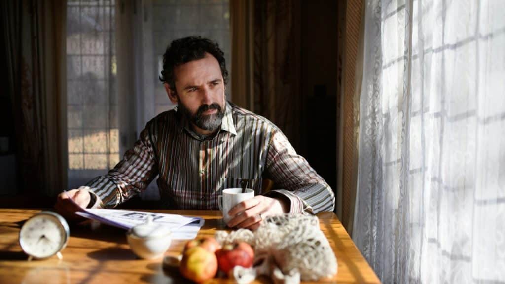 A bearded man in a striped shirt sits at a sunlit table with a mug and newspaper, looking out a window.