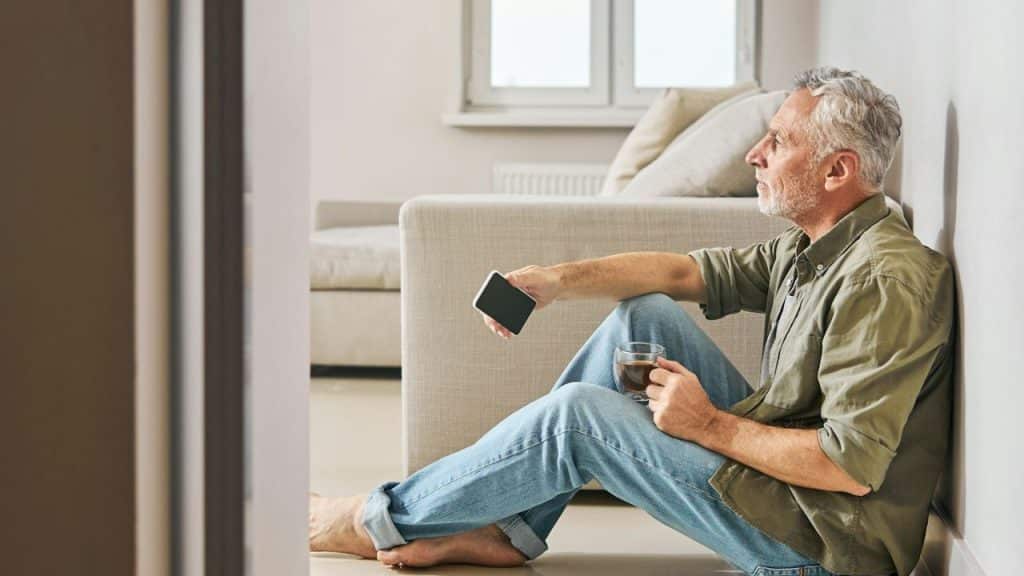 Older man sitting on the floor with a phone and drink, looking away.