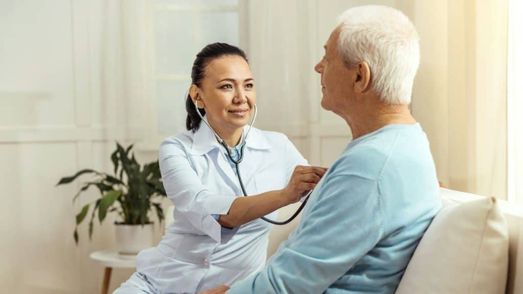 Female doctor in a white coat listening to an older man's chest with a stethoscope.