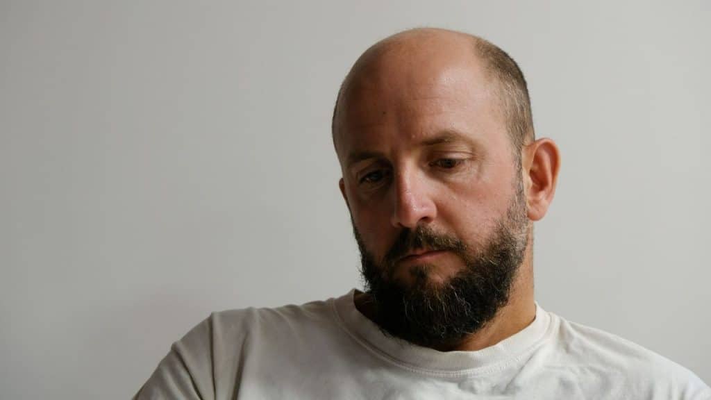 Close-up of a balding man with a dark beard looking down against a light background.