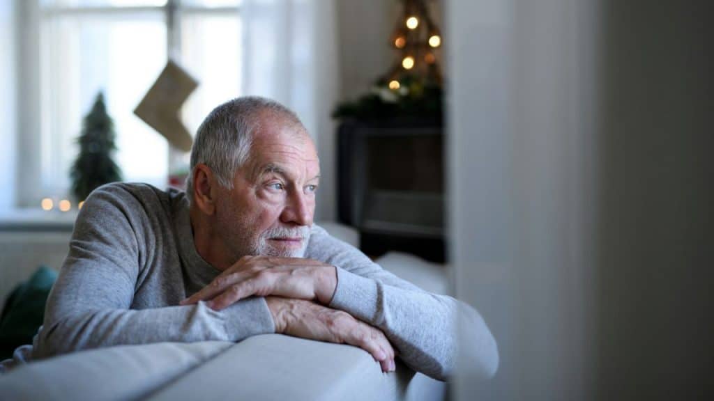 Senior man with a gray beard resting on a couch, looking away in a dim room.